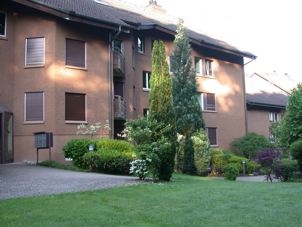 A multi-story residential building with a brown facade, multiple windows, balconies, and surrounding greenery including trees and shrubs.