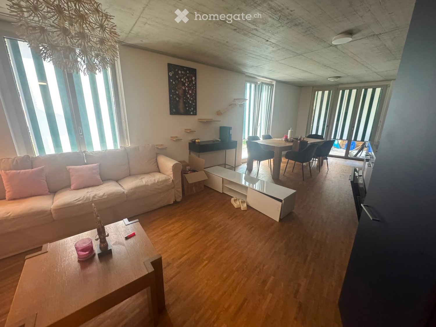 Modern living room with a white sofa, wooden coffee table, dining area with chairs and table, and large windows with blinds.
