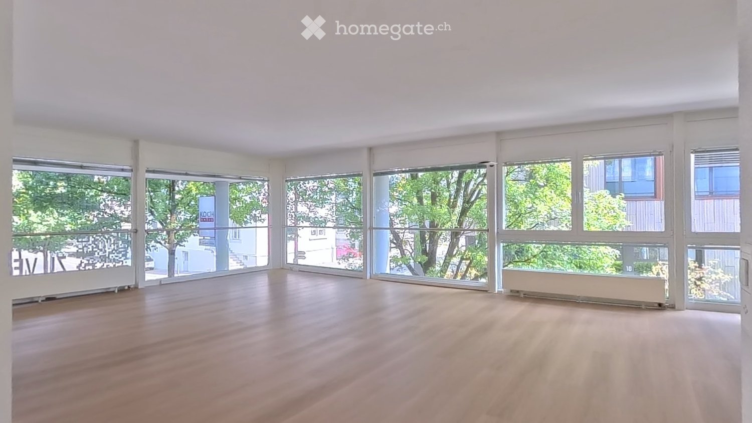 Empty room with wooden flooring and large windows offering a view of greenery outside. No furniture or equipment present.
