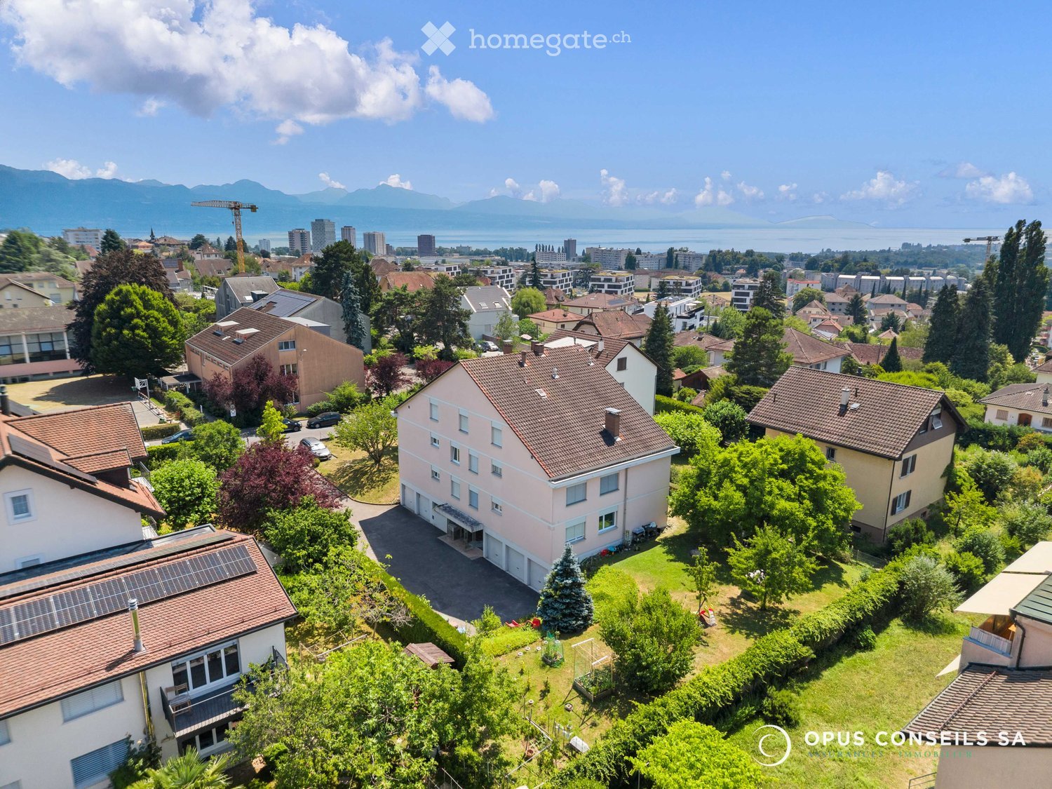 Aerial view of a suburban neighborhood with multiple houses, gardens, and a scenic backdrop of mountains and a lake.