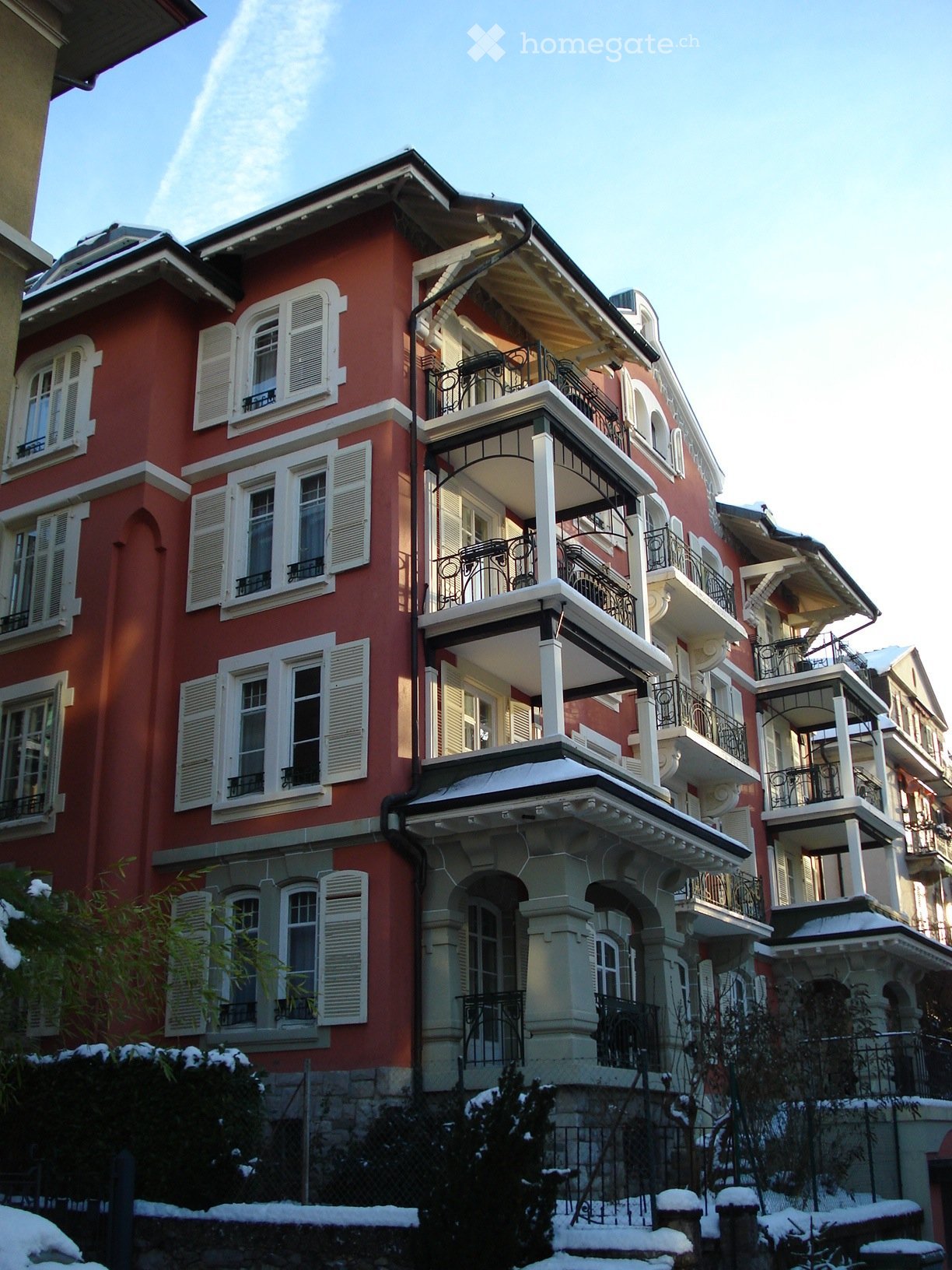 A multi-story red brick building with white-framed windows and balconies, featuring ornate railings and a snow-covered garden area in front.