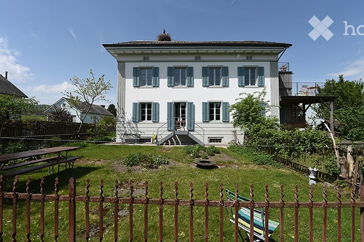 A well-maintained garden area in front of a two-story house with white walls and blue shutters. The garden features a wooden fence, a picnic table, and a small seating area with a bench and chairs.