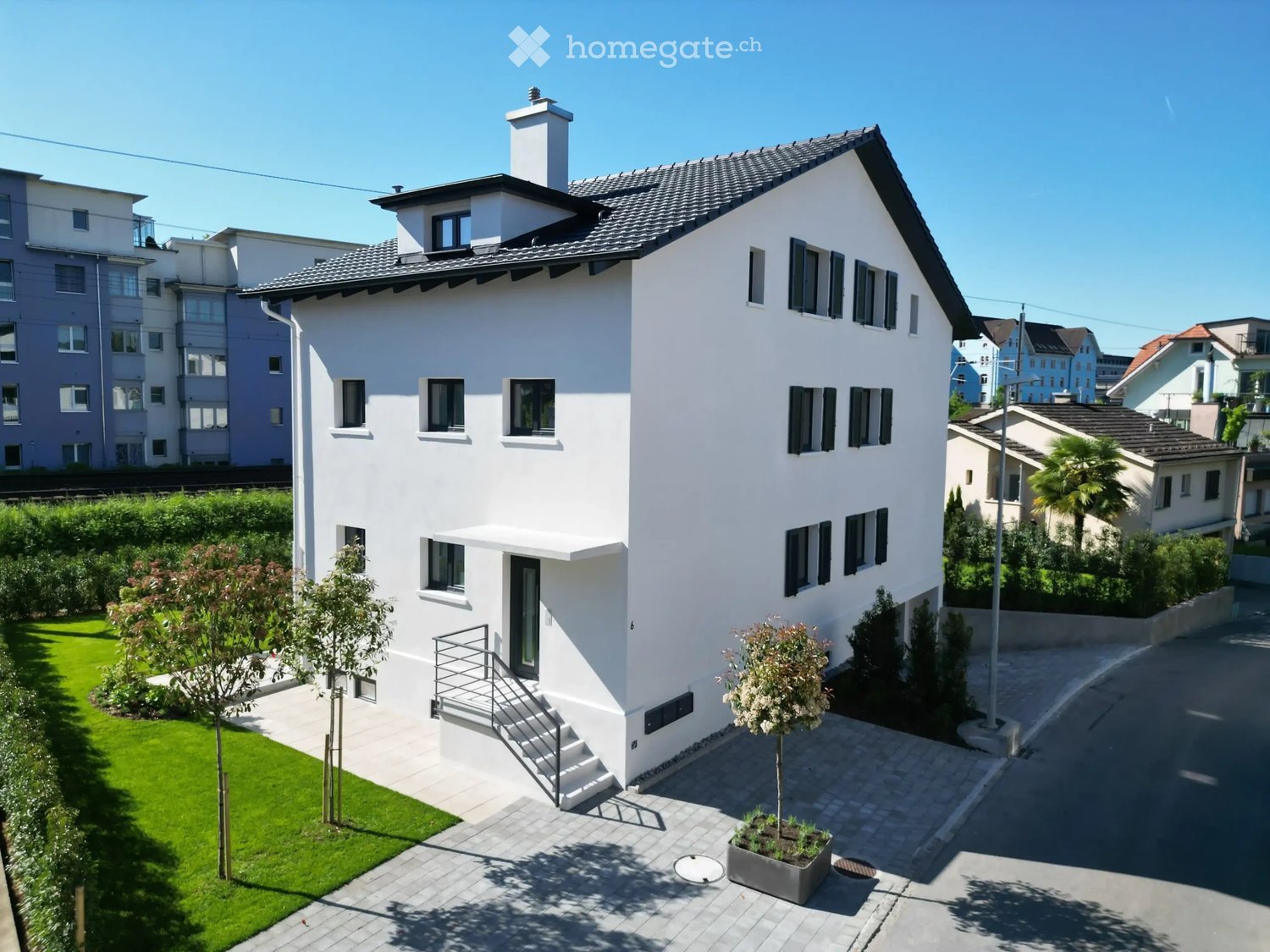 Modern two-story white house with a dark roof, multiple windows, and a small staircase leading to the entrance. The house is surrounded by a well-maintained lawn, small trees, and a paved pathway.