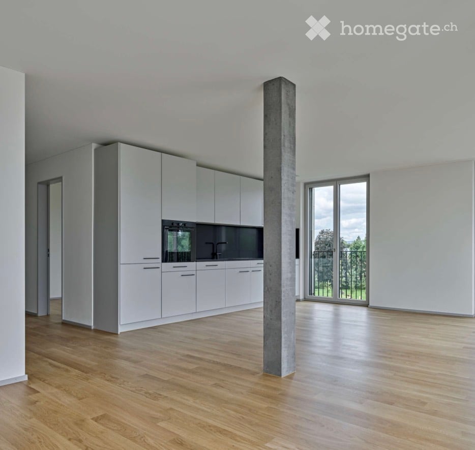 Empty kitchen with white cabinets, wooden flooring, and a large window overlooking a green area.