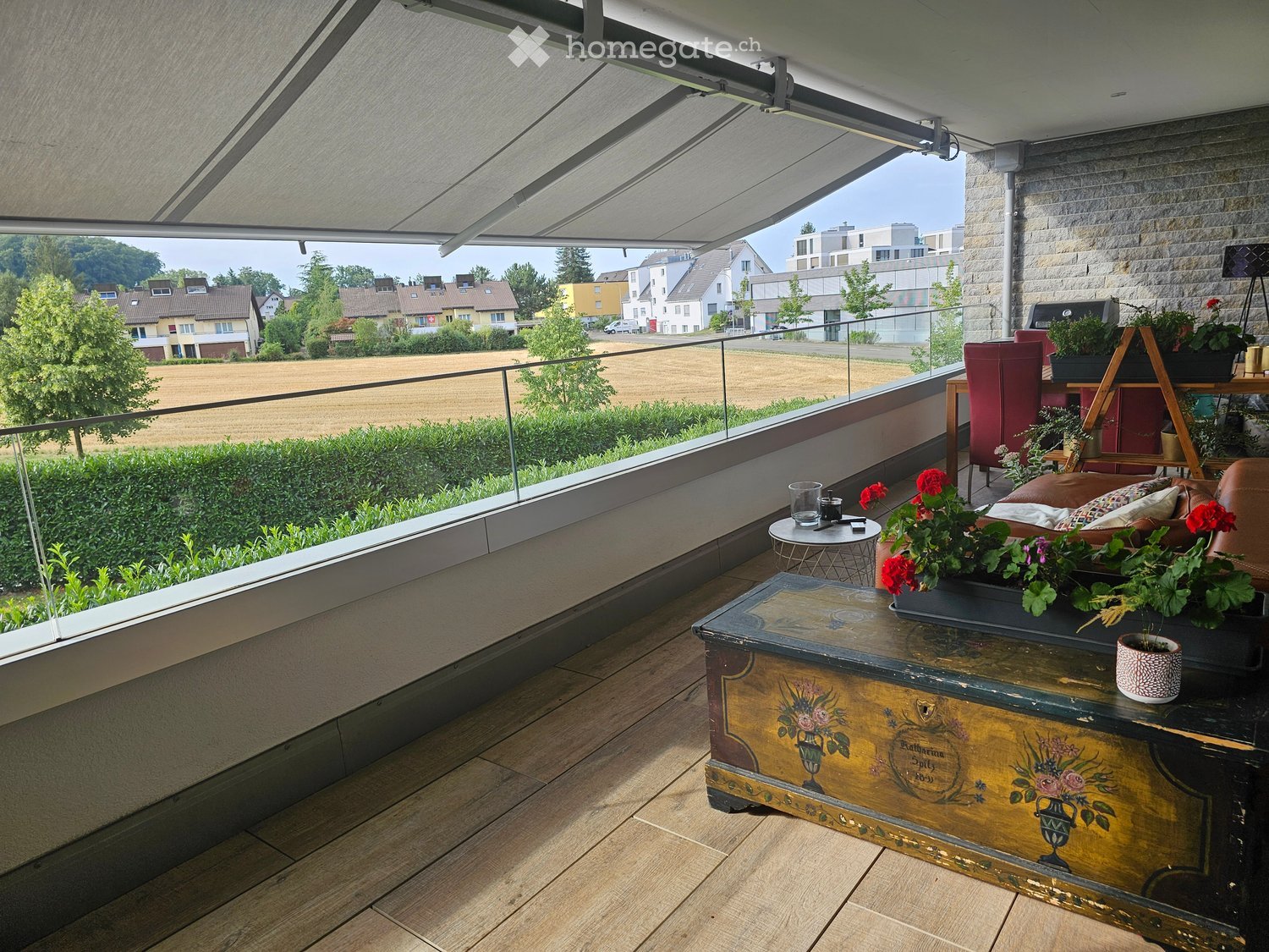 Balcony with wooden decking, glass railing, sofa, coffee table, flower pots, and a view of the countryside.