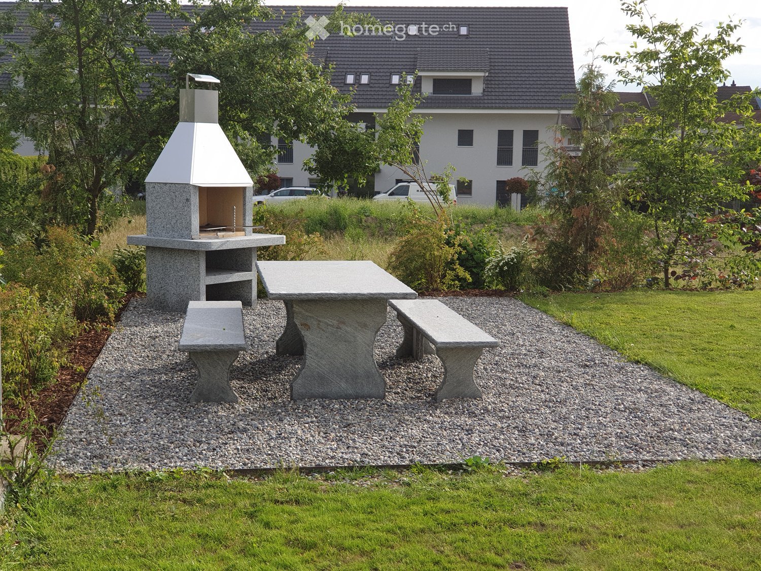 Garden area with a stone barbecue, stone table, and benches on a gravel path, surrounded by greenery and a house in the background.