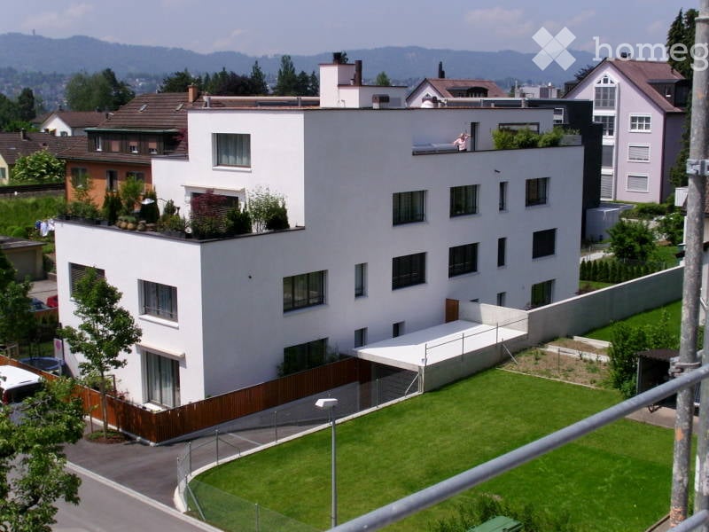 Modern white multi-story building with flat roof, large windows, and rooftop greenery. Surrounded by other houses and greenery with a scenic mountain view in the background.