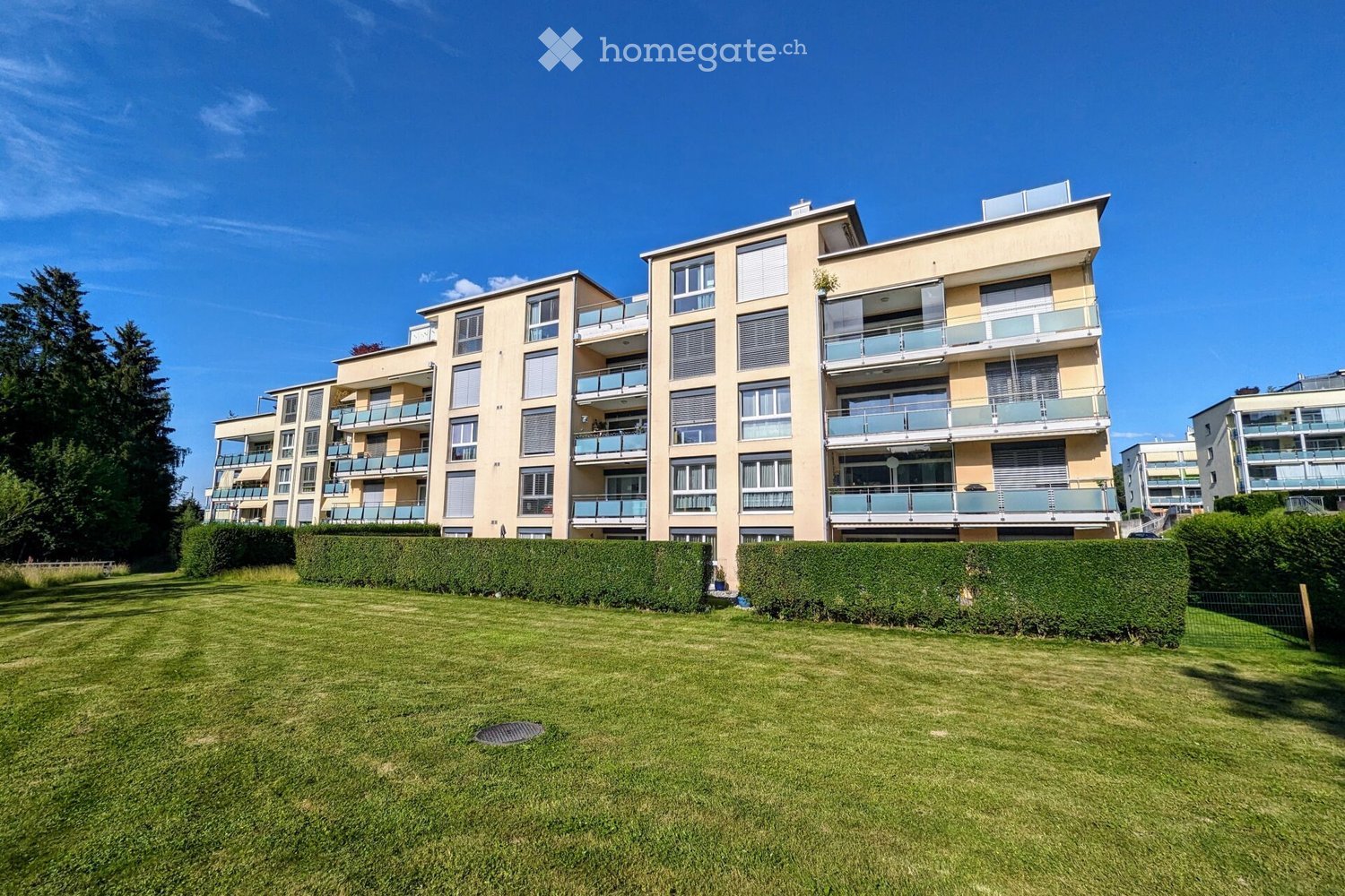 Modern multi-story residential building with balconies, surrounded by a well-maintained lawn and hedges.