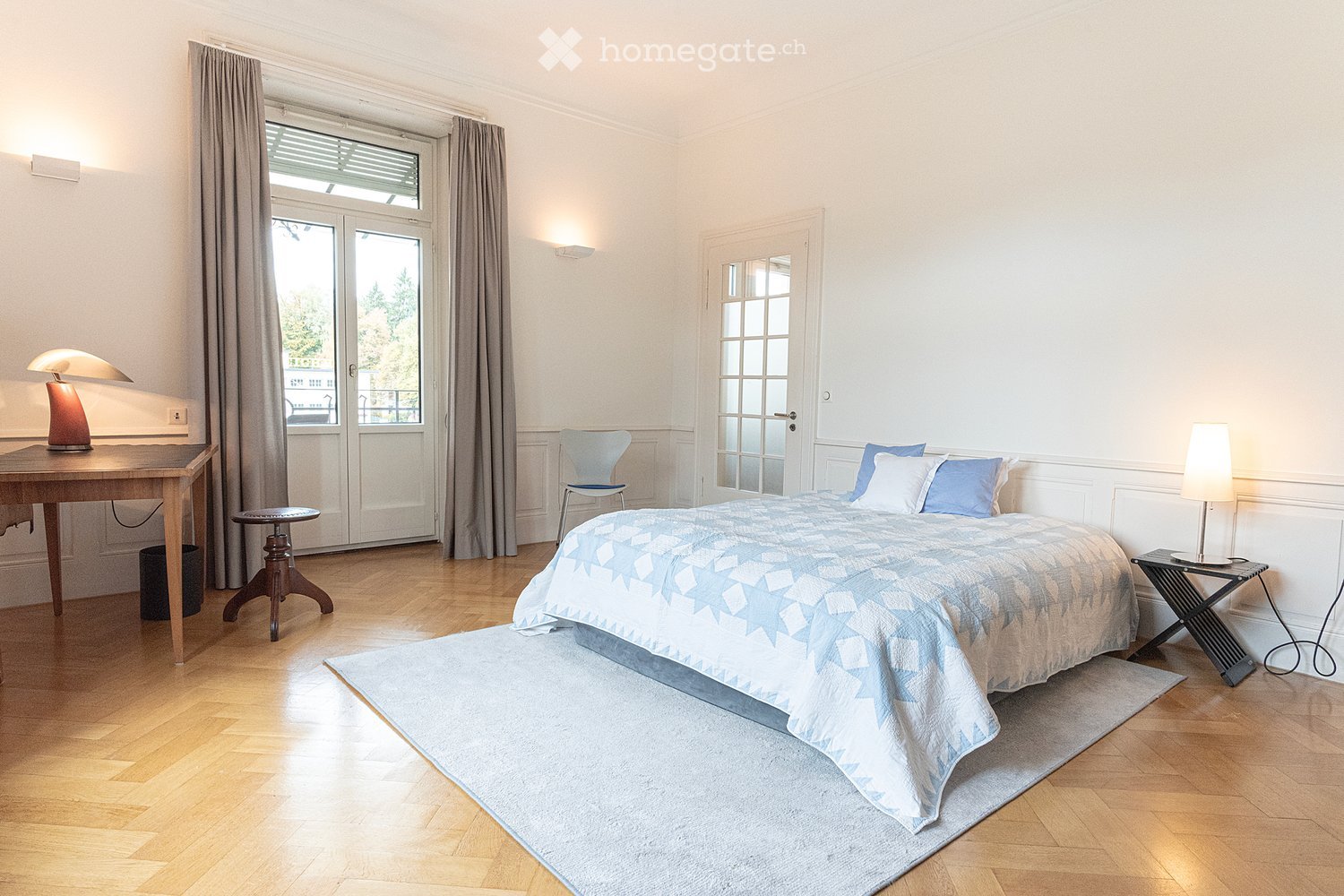 Bright bedroom with wooden flooring, large bed with blue and white bedding, nightstand, desk with lamp, chair, and a small stool near the window.
