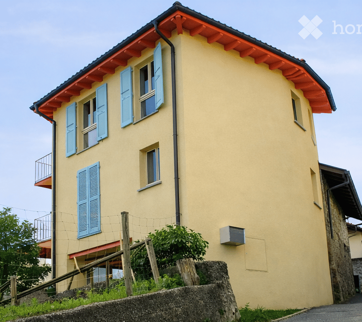 Two-story house with yellow walls, blue shutters, and a red-tiled roof. Features a small balcony on the upper floor and a stone retaining wall in front.