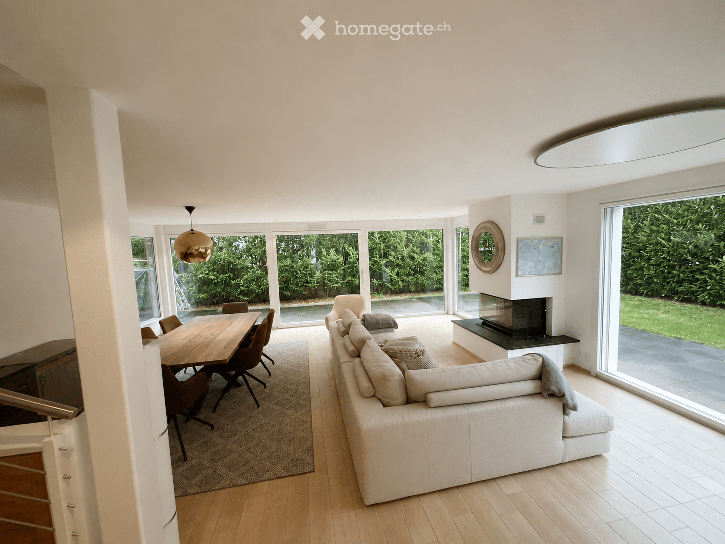Bright living room with large windows and a view of greenery, featuring a white sectional sofa, a wooden dining table with chairs, a fireplace, and modern decor including a pendant light and artwork.