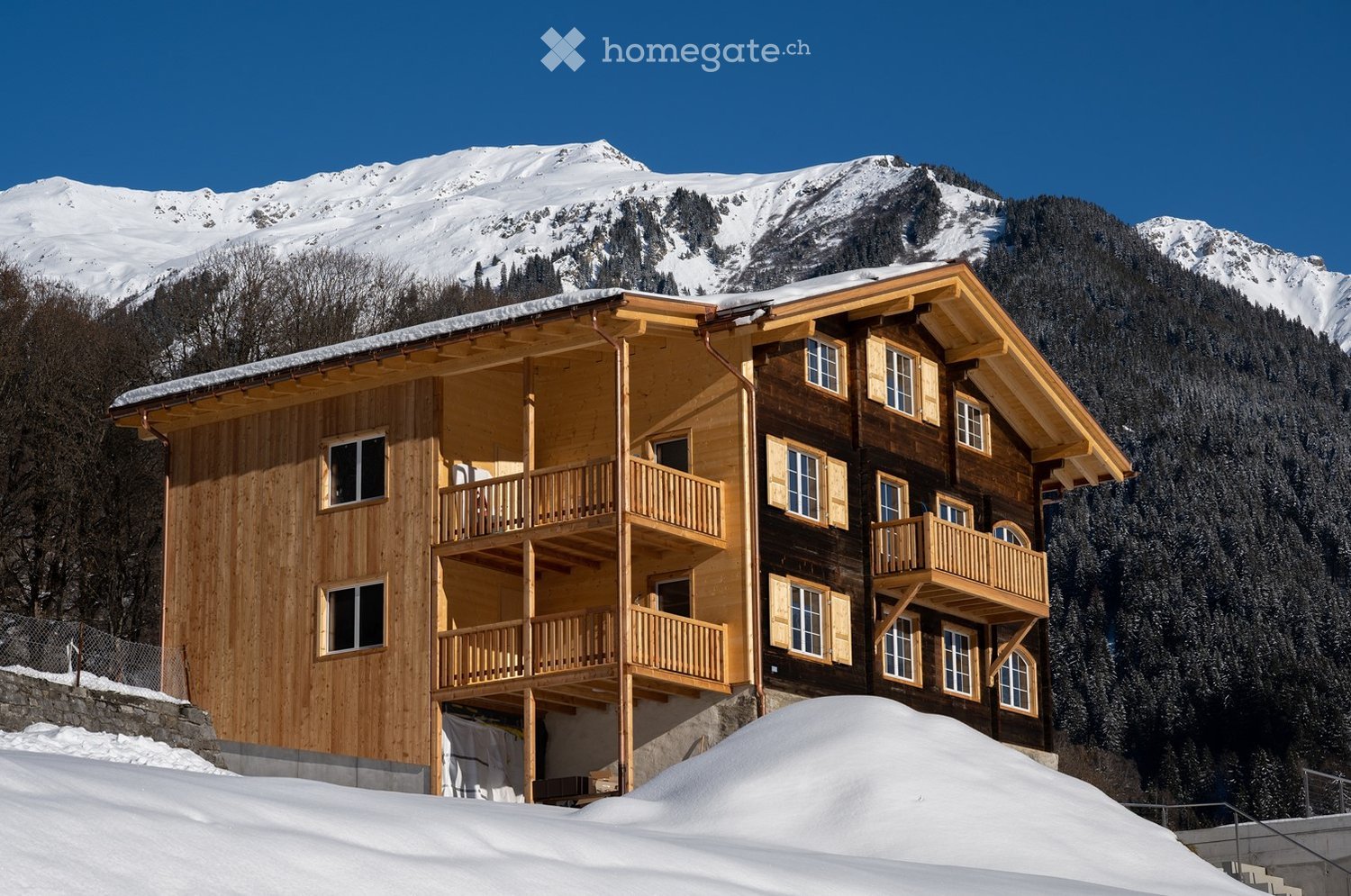 A large, multi-story wooden chalet with balconies and large windows, set against a backdrop of snow-capped mountains and a clear blue sky.