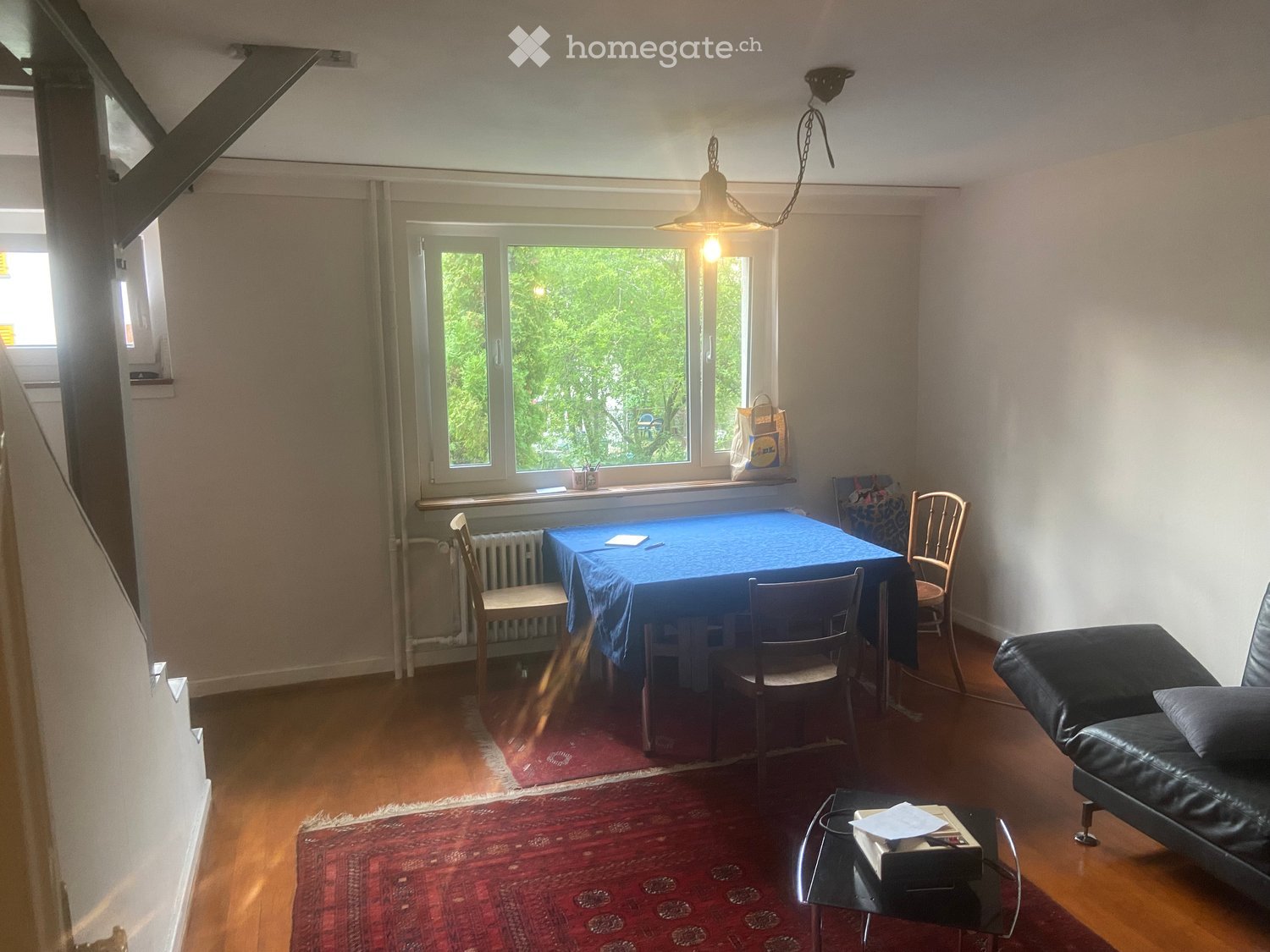 A cozy living room with wooden flooring, a large window, a sofa, a coffee table, a dining table with chairs, and a hanging light fixture.