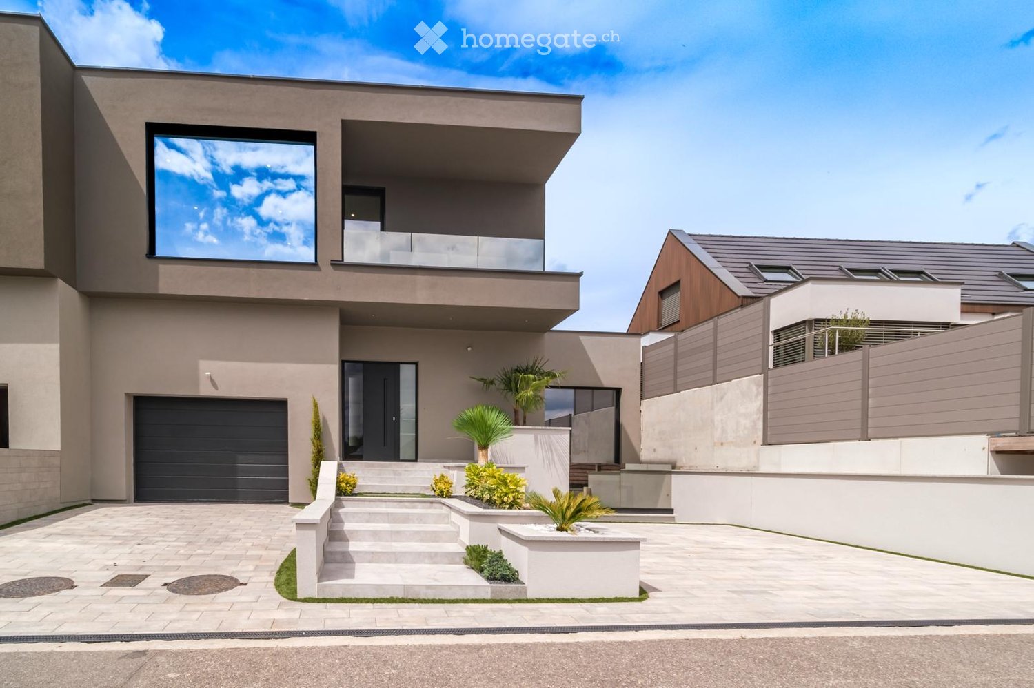 Modern two-story house with a sleek design, featuring a large garage, balcony, and contemporary landscaping with small plants and a raised garden bed.