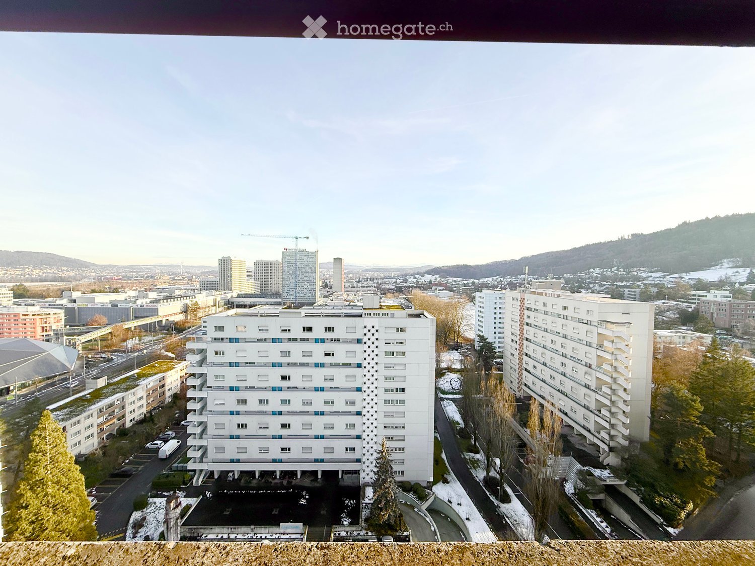 A multi-story residential building with white facade and blue accents, surrounded by other buildings and a parking lot, with snow-covered ground and distant cityscape.