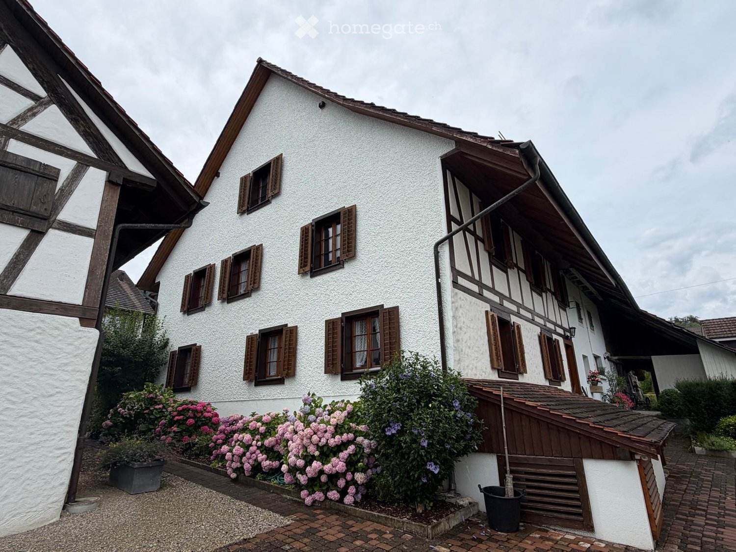 Traditional two-story house with white plaster walls, wooden shutters, and a wooden balcony on the upper floor. The house features a pitched roof and is surrounded by a garden with pink flowers and shrubs.