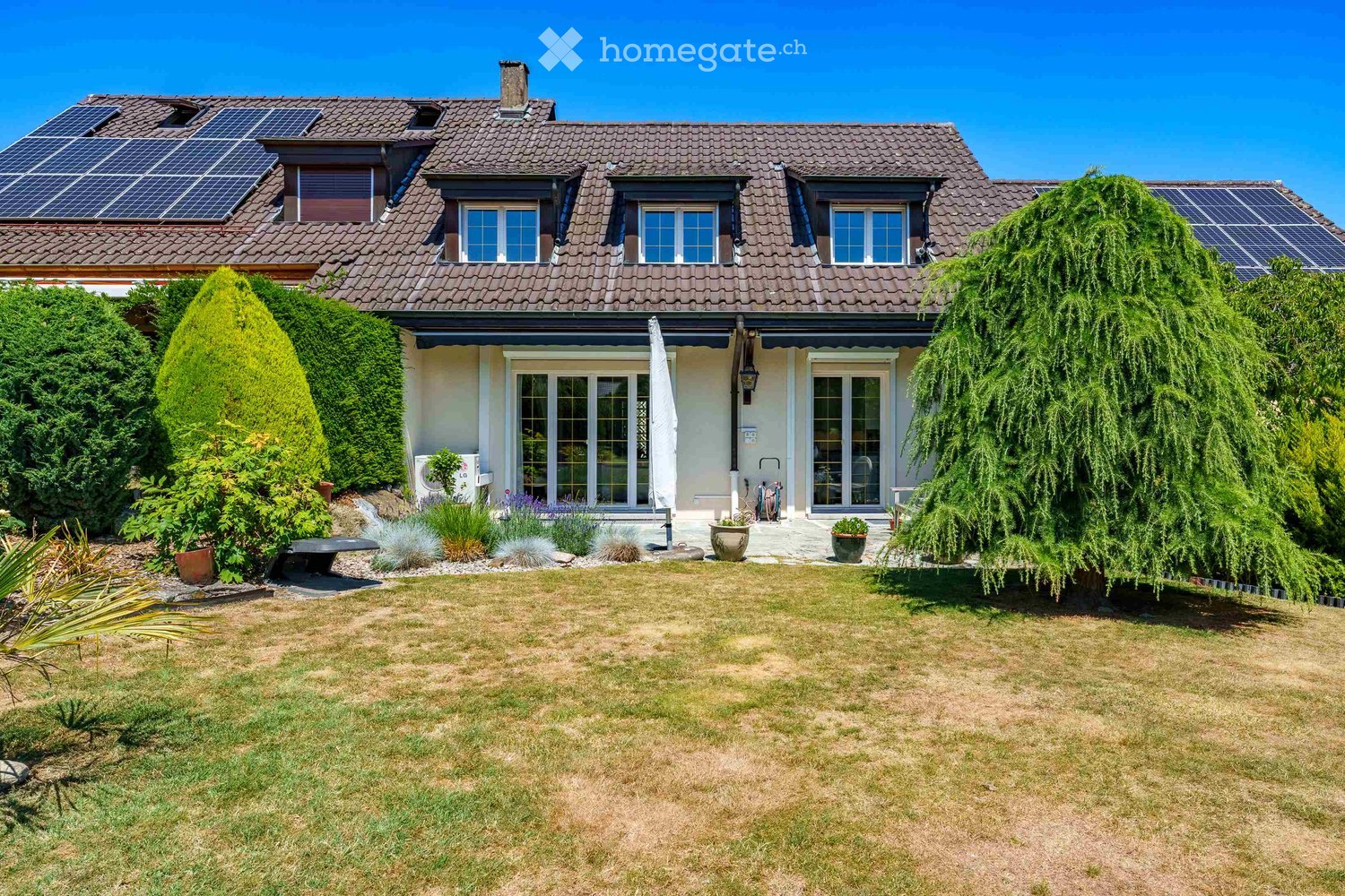 Two-story house with a tiled roof, solar panels, large windows, and a well-maintained garden with a lawn, shrubs, and potted plants.