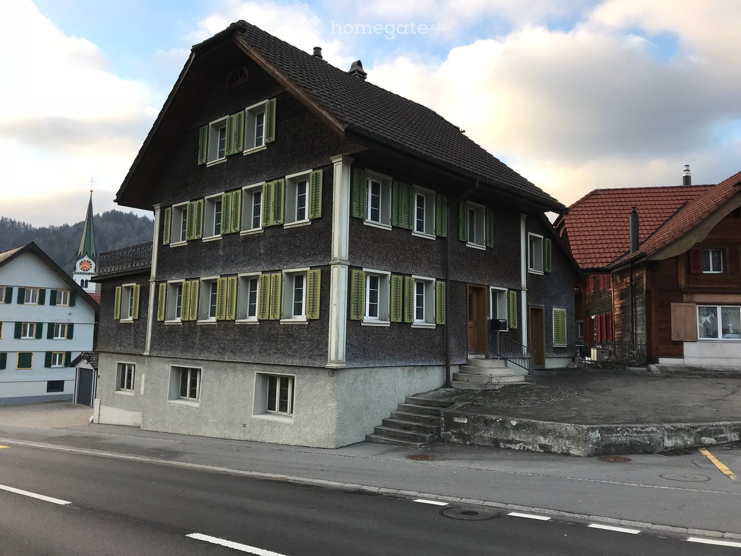 A two-story building with a dark roof and green shutters, situated on a street with a church visible in the background.