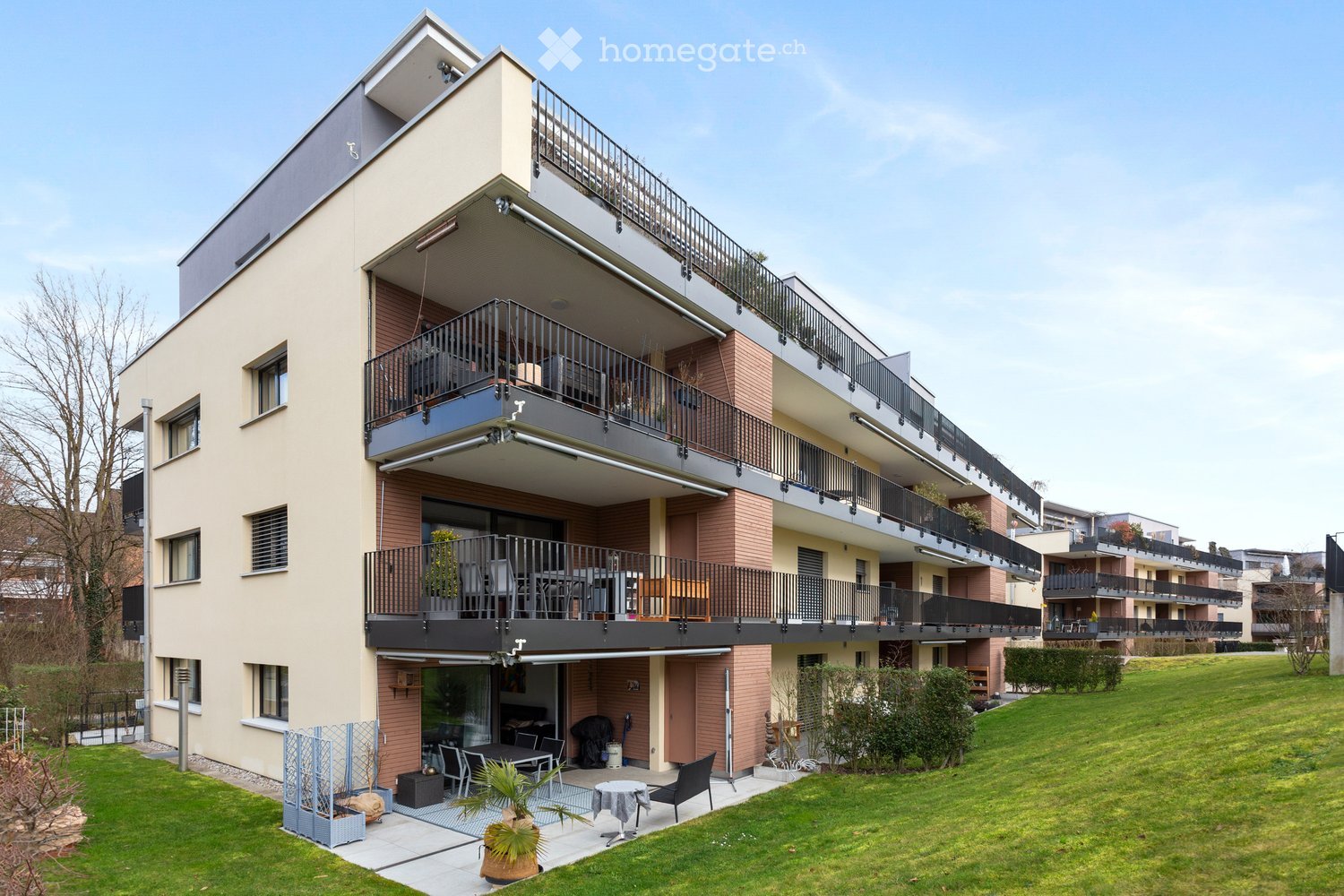 Modern multi-story residential building with balconies, large windows, and a well-maintained lawn area with outdoor furniture.