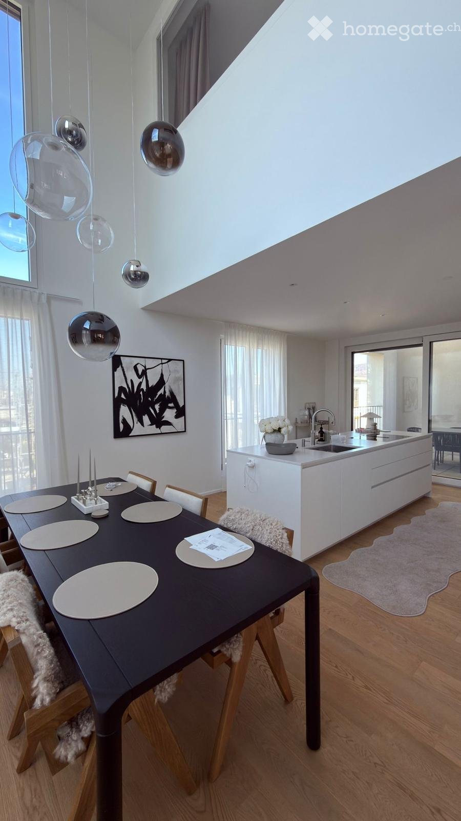 Modern dining area with a dark wooden dining table, white chairs, pendant lights, and a white kitchen island with a sink and faucet. A vase with white flowers sits on the island, and a picture frame hangs on the wall.