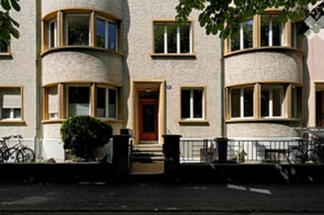 Raised ground floor, two-story building, beige exterior with yellow window frames, multiple arched windows, two bicycles parked in front