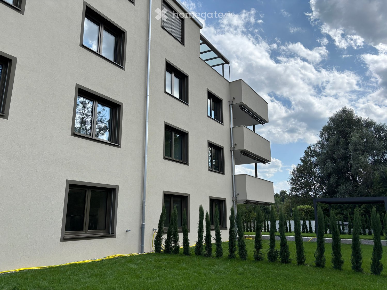 Modern multi-story building with large windows, balconies, and surrounding greenery.