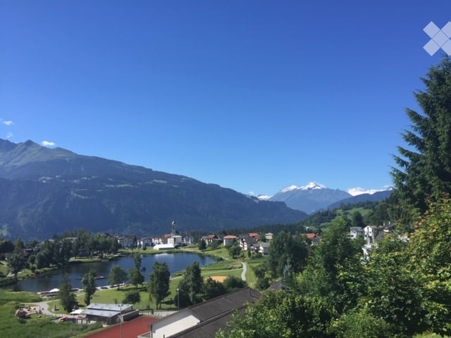 Scenic outdoor view of a town nestled in a valley surrounded by mountains, with a lake and lush greenery.