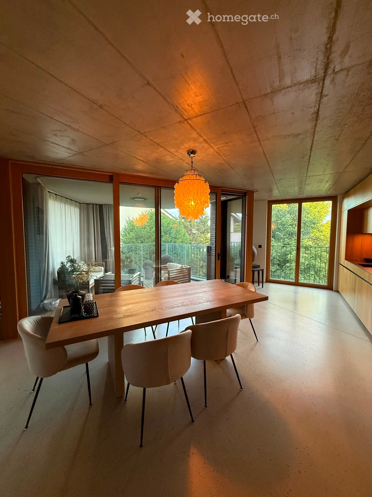 Modern dining room with a long wooden dining table, six chairs, a hanging ball lamp, large windows with curtains, and a view of a balcony area with outdoor furniture.