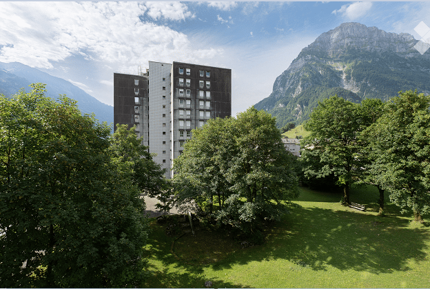 A modern apartment building surrounded by lush green trees and a scenic mountain backdrop. The building has multiple floors with numerous windows and is situated in a serene, green landscape.