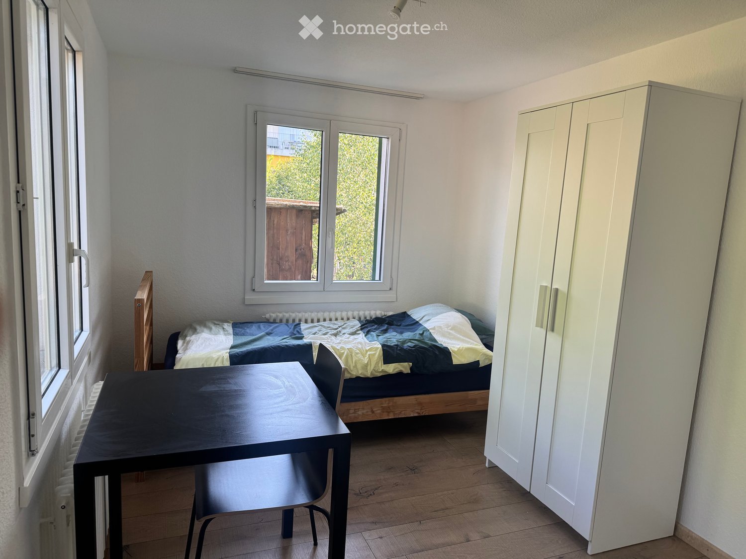 A small, furnished bedroom with a bed, a table, a chair, and a wardrobe. The room has wooden flooring and large windows with a view of greenery.