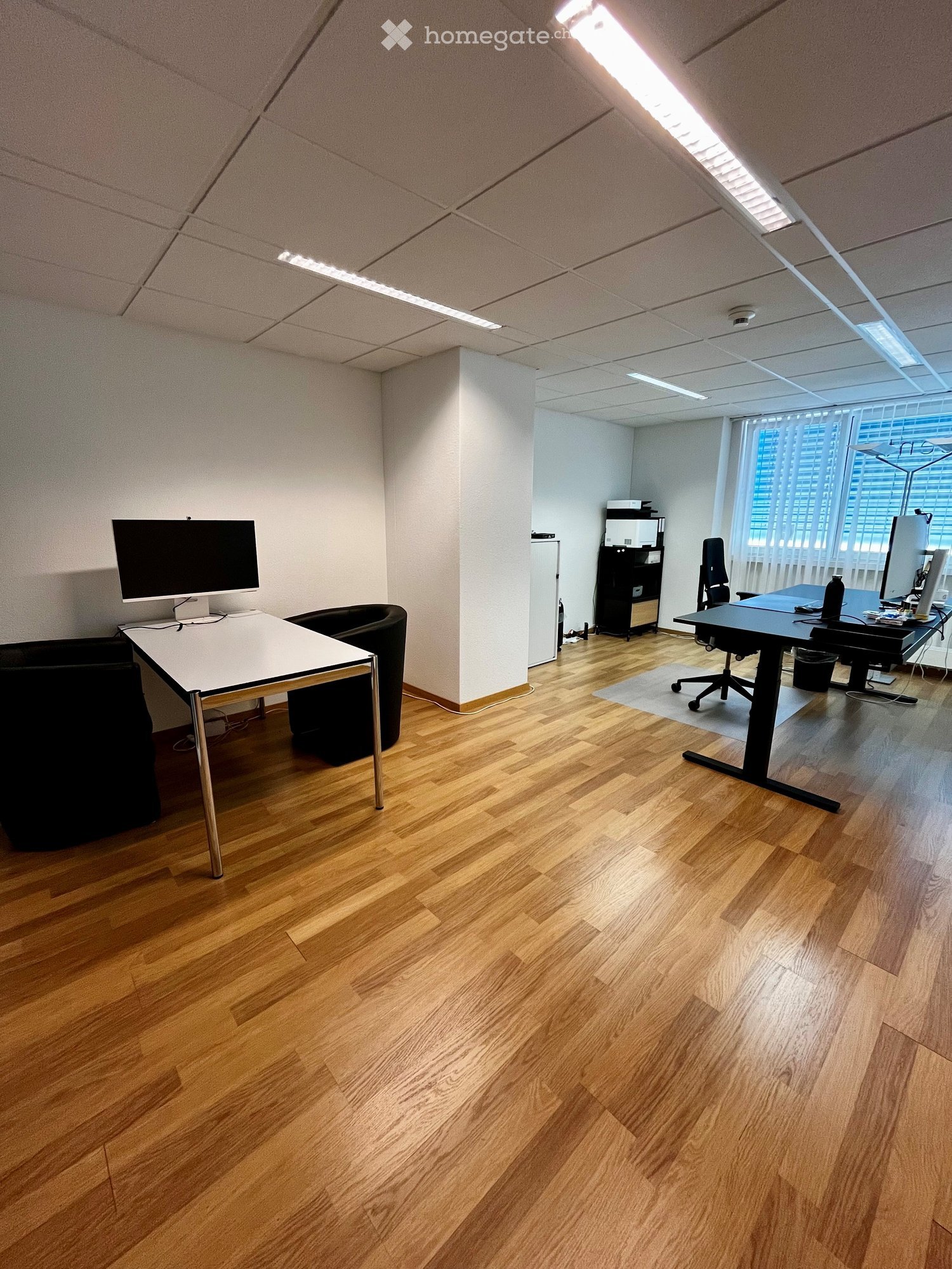 Office space with wooden flooring, two desks, chairs, and computer screens. Features modern lighting and large windows with blinds.