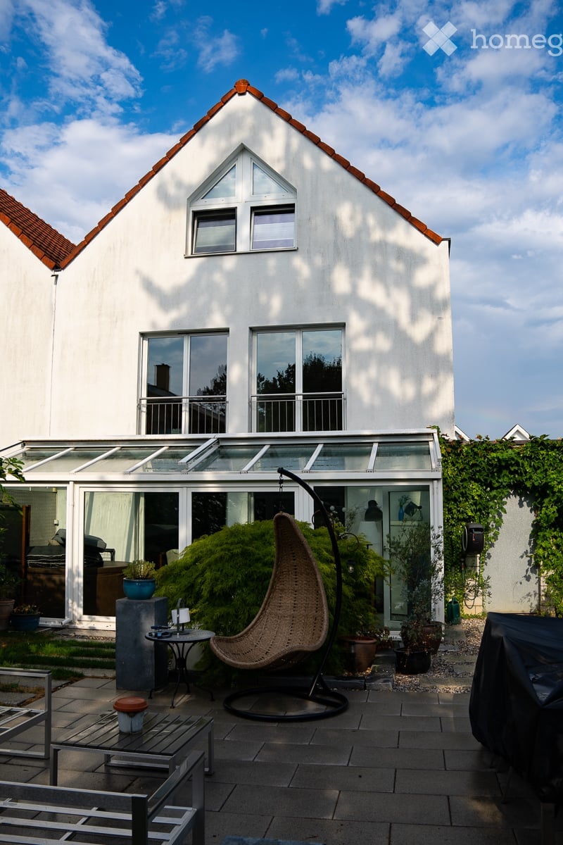 A modern patio area with a wicker hanging chair, small table, and several potted plants. The patio is attached to a white house with large windows and a glass roofed extension.