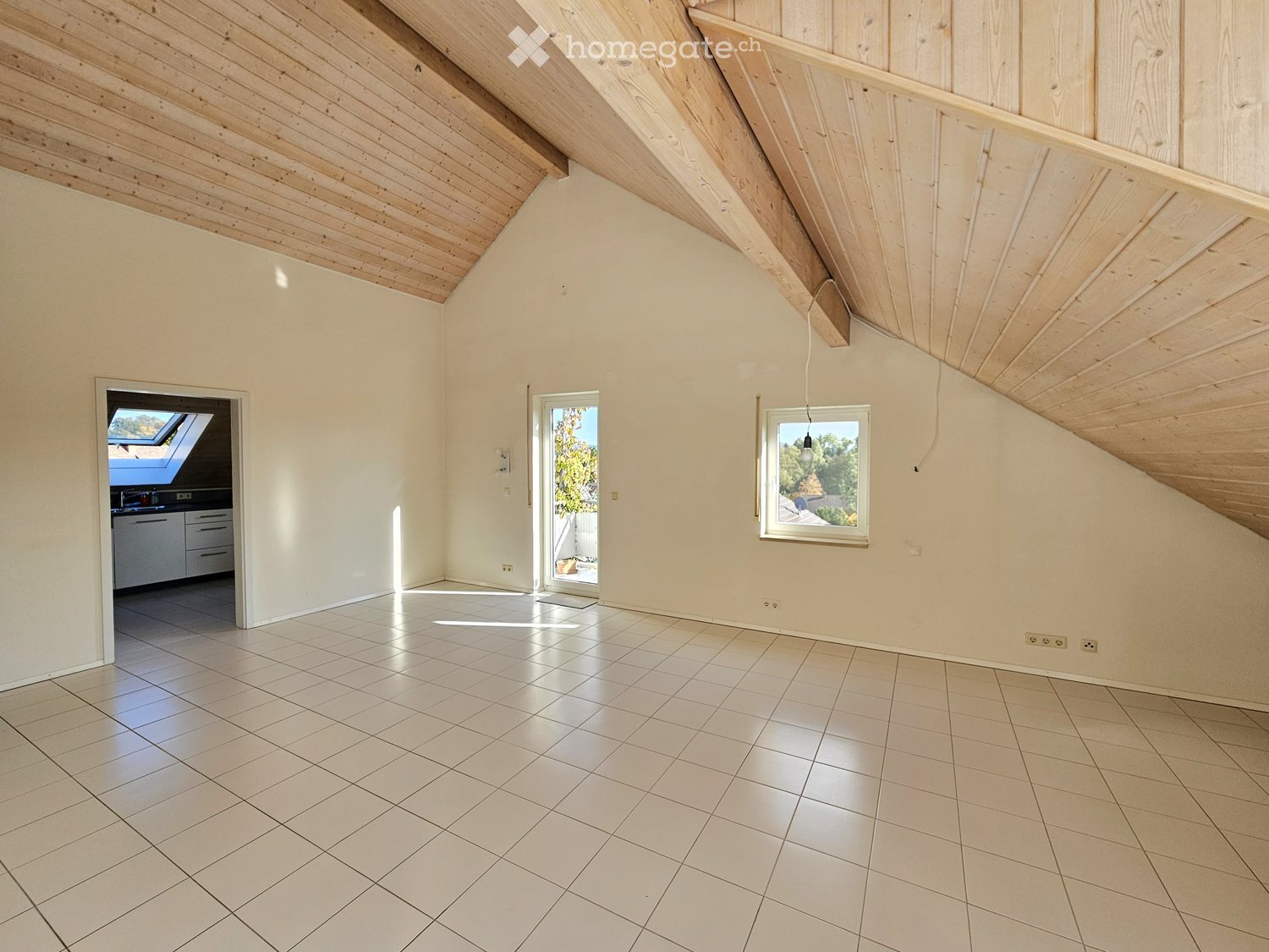 Bright, empty room with white tiled flooring, wooden slanted ceiling, and large windows allowing natural light. Features an open doorway leading to another room and a skylight above.