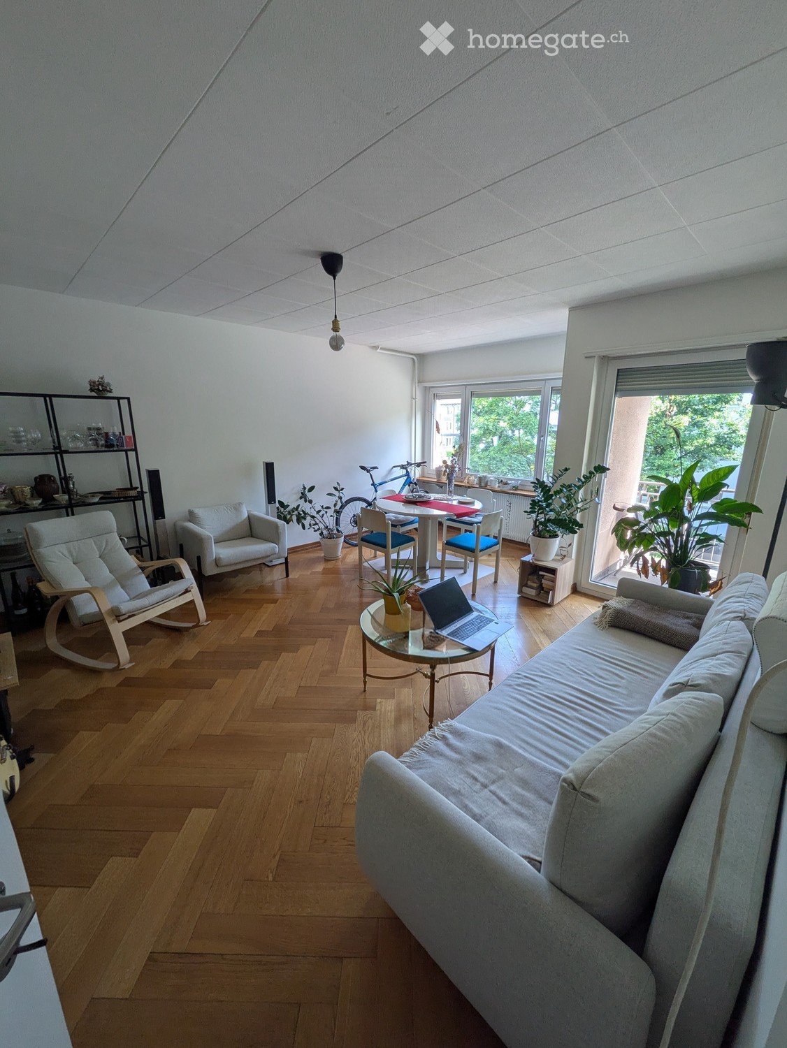 Bright living room with wooden flooring, white walls, and large windows. Features a grey sofa, armchair, coffee table with laptop, dining table with chairs, and shelves with decorative items.