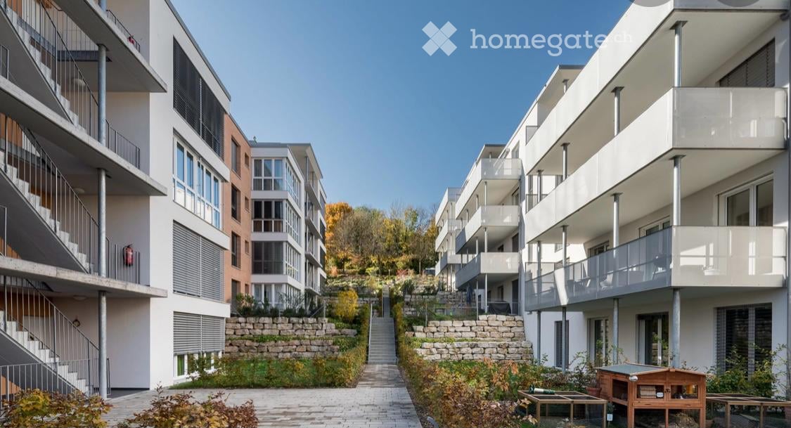 Modern multi-story residential building with balconies, raised ground floor, and landscaped garden area.
