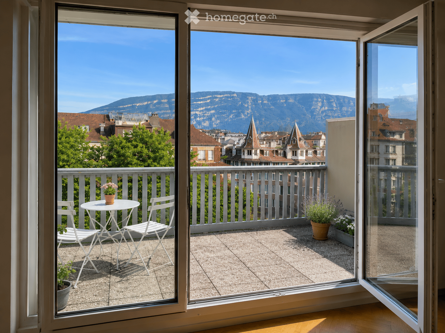 Balcony with a view of city rooftops and mountains, featuring a small table, two chairs, and potted plants.