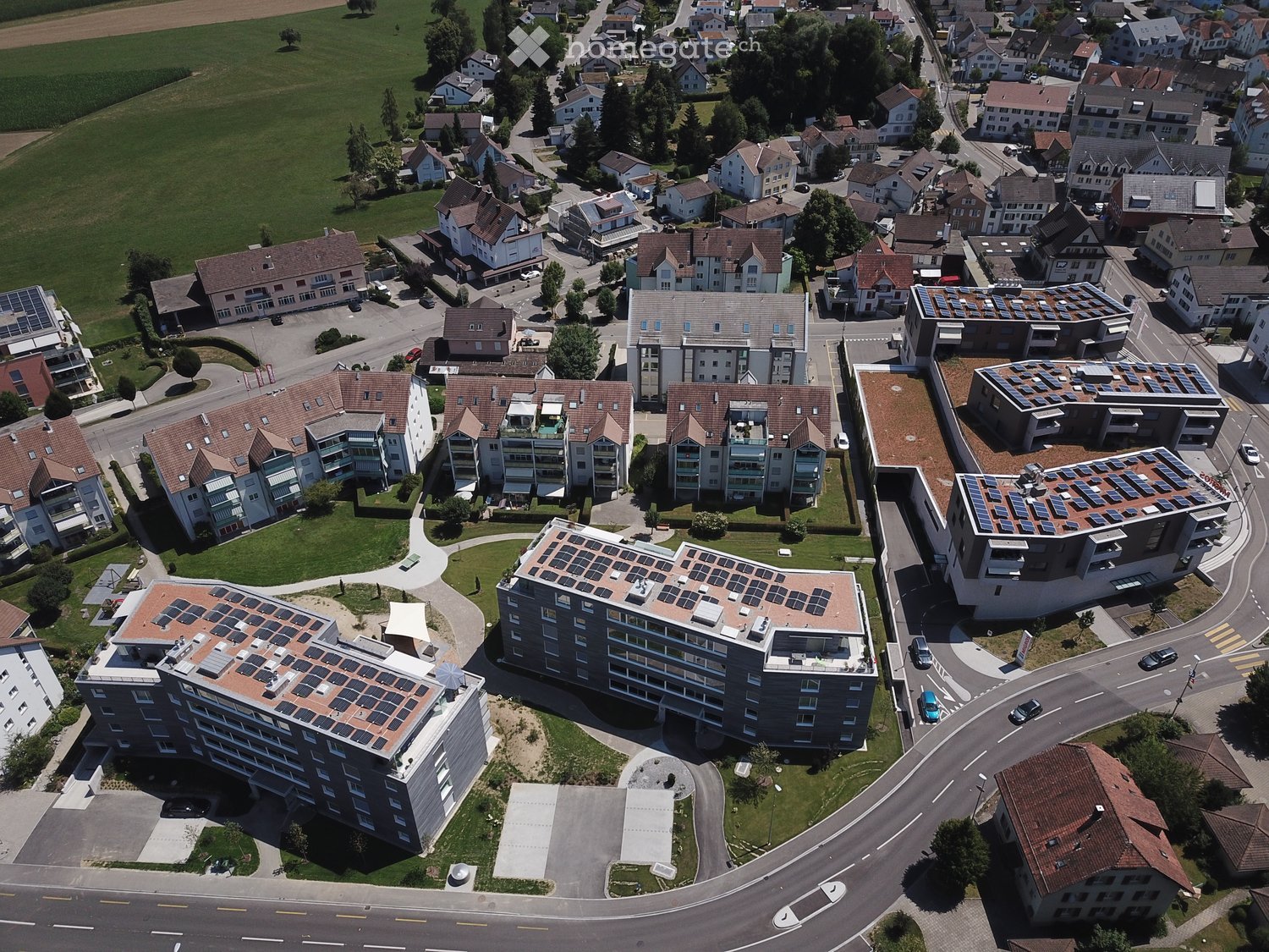 Aerial view of a residential area with multiple buildings, including apartment complexes with solar panels on the roofs, surrounded by green spaces and roads.