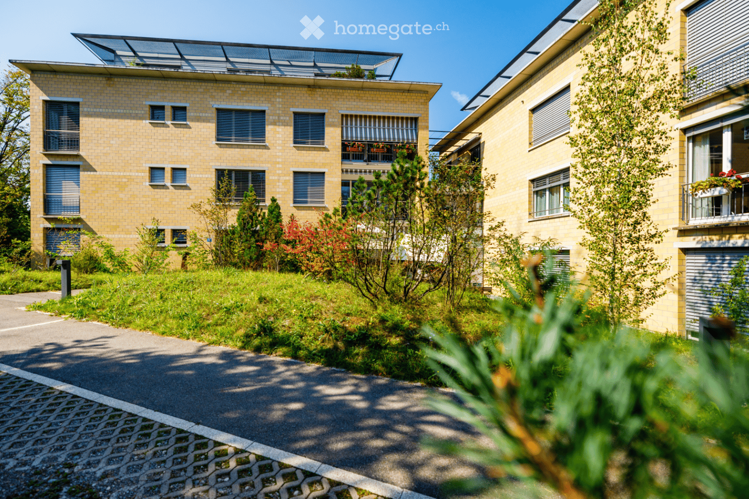 Two-story residential building with yellow brick facade, multiple windows, and a rooftop terrace. The building is surrounded by greenery and a paved pathway.