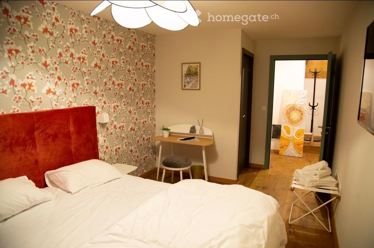 Cozy bedroom with a red headboard bed, a small wooden desk with a stool, a picture frame on the wall, and a plant on the desk. A hallway leads to another room with a colorful tapestry.