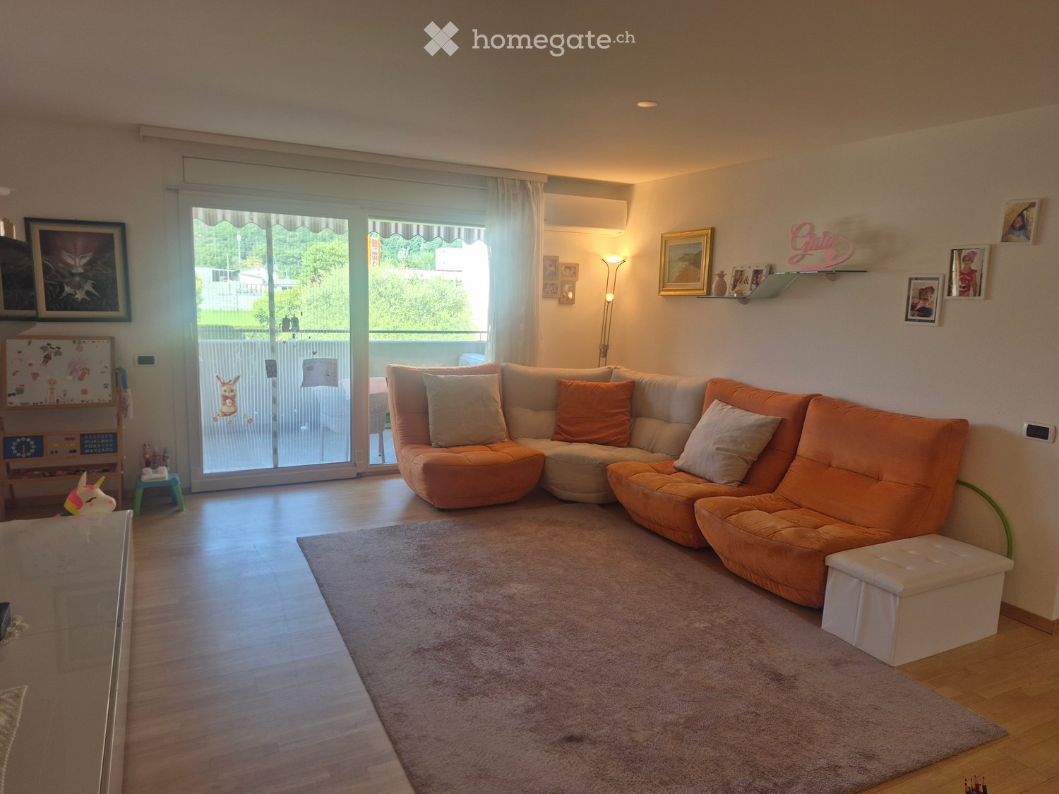 Modern living room with a large orange sectional sofa, a glass coffee table, a floor lamp, and a shelf with picture frames and decorative items. The room features wooden flooring, a large window with a view, and a small child's play area in the corner.