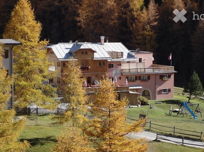 A multi-story building with a mix of wooden and pink facades, surrounded by autumn-colored trees and a fenced yard with playground equipment.