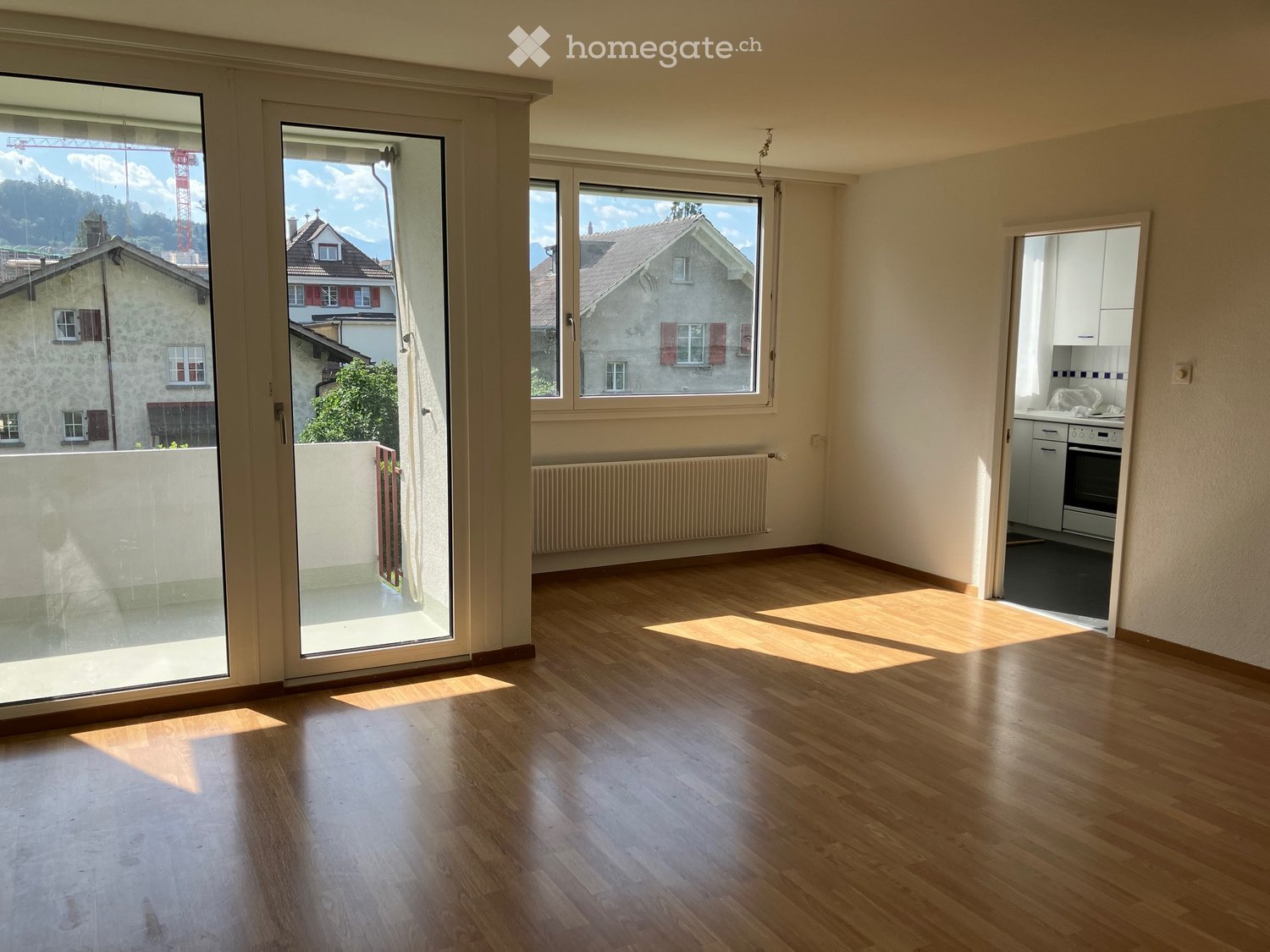 Bright, unfurnished living space with wooden flooring, large windows, and a view of the surrounding area. Features a sliding glass door leading to a balcony.