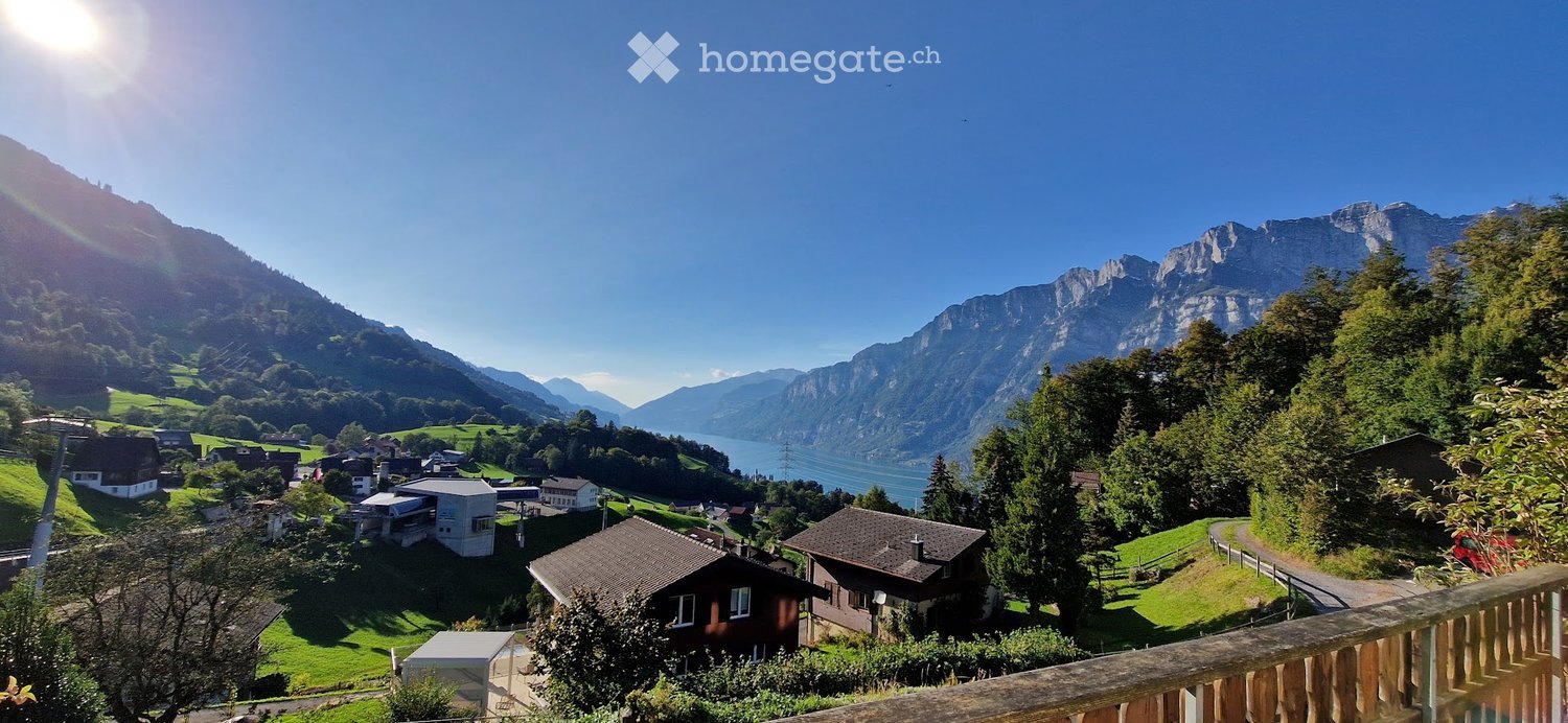 Panoramic view of a Swiss village nestled in a valley surrounded by mountains and a lake, with houses and lush greenery visible.