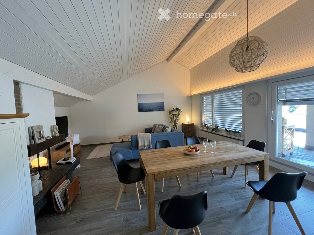 Modern living and dining area with wooden flooring, white walls, and a slanted ceiling. Features a blue sofa, wooden dining table with chairs, a small side table, and decorative items including a picture frame and a clock.