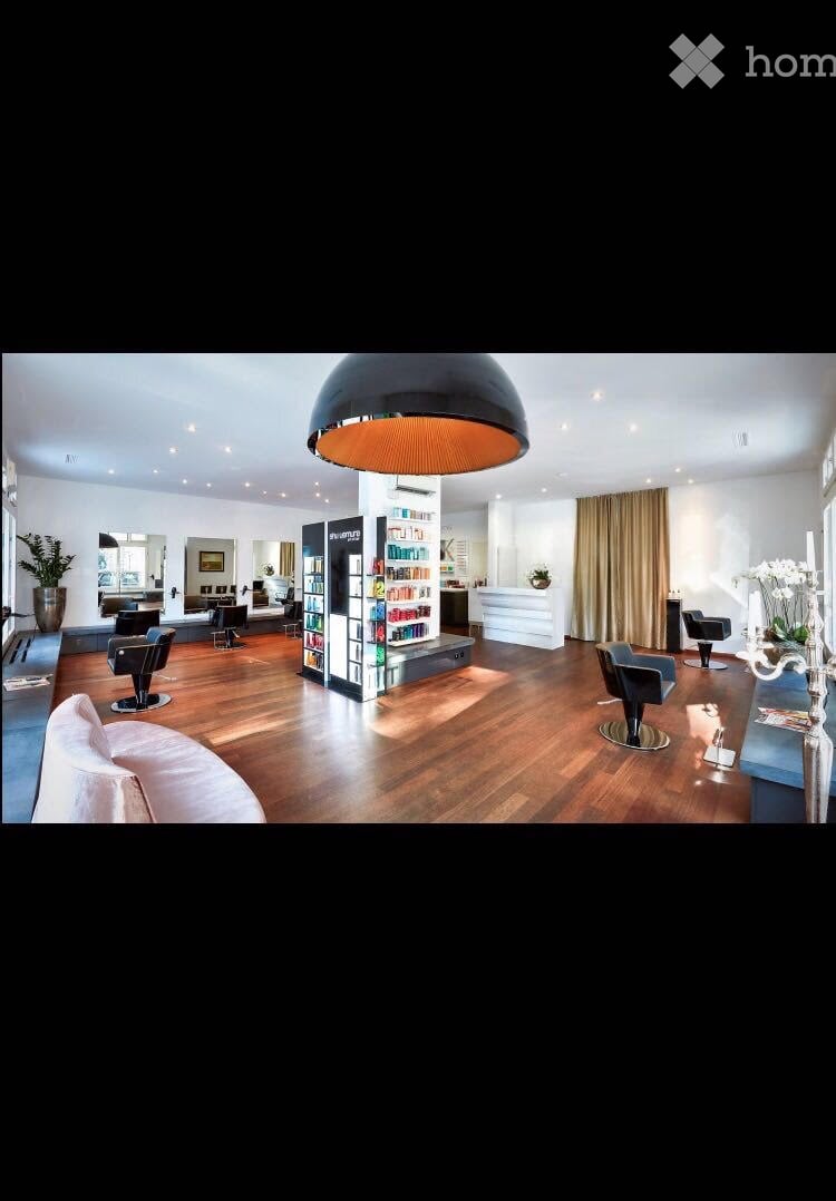Modern, spacious salon with wooden flooring, large round pendant light, several chairs, a sofa, shelves with products, and a reception desk.