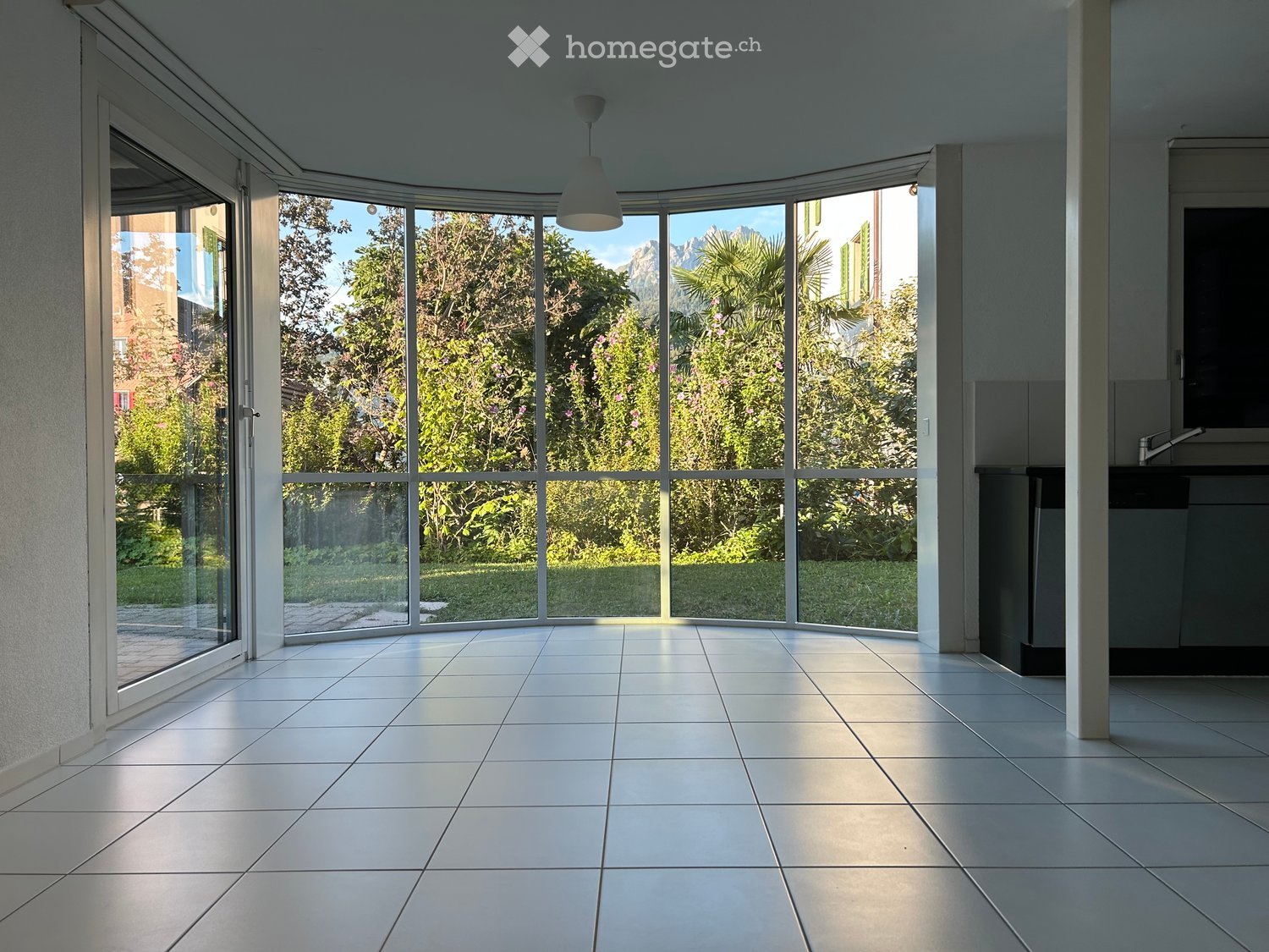 Bright, empty room with large glass windows and doors, tiled floor, and modern lighting fixture.