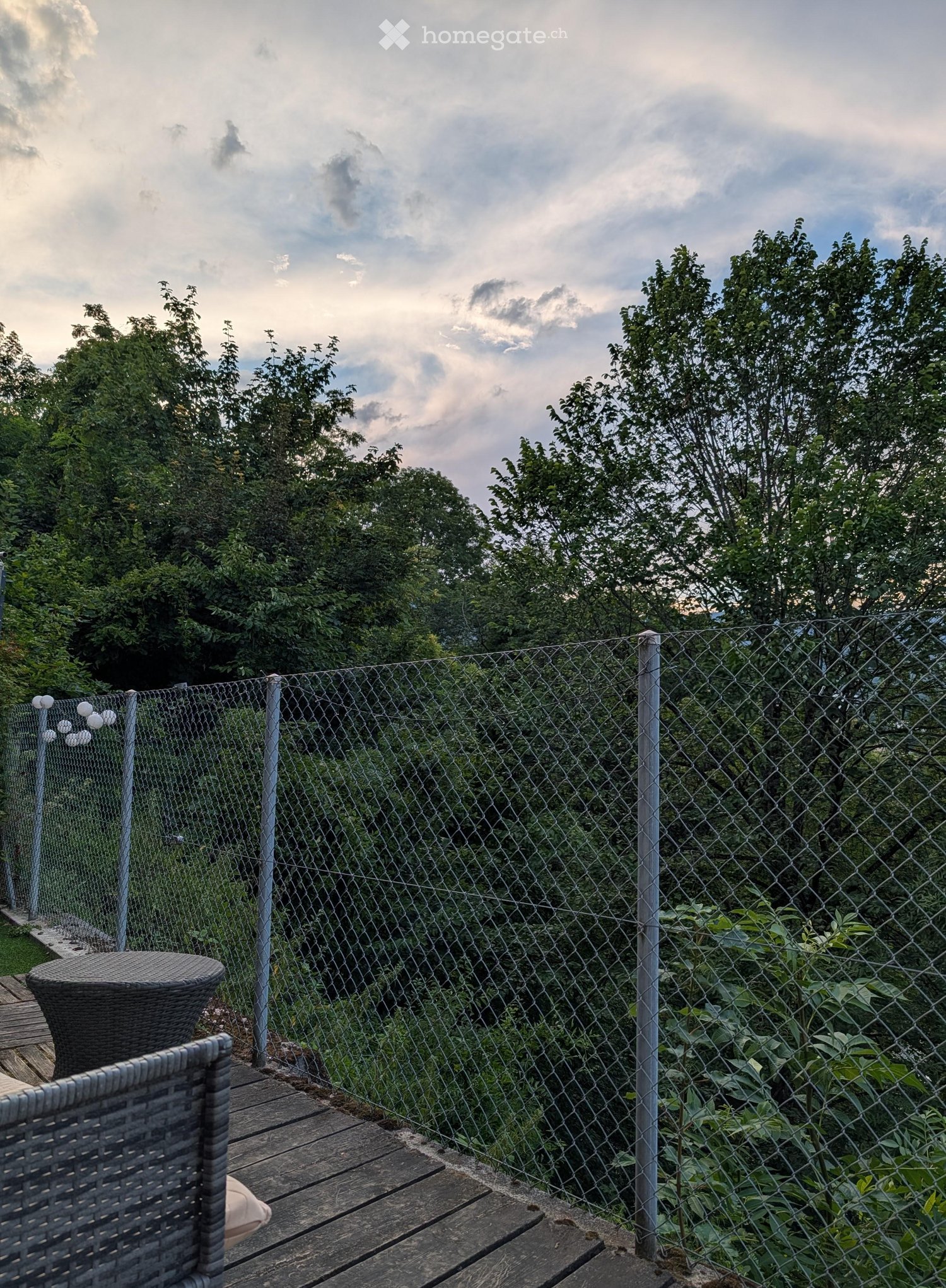 A wooden balcony with a wicker chair and table, enclosed by a chain-link fence, overlooking a lush green area with trees.