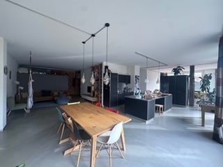 Open-plan living and dining area with modern kitchen, wooden dining table, pendant lights, and a couch in the background.