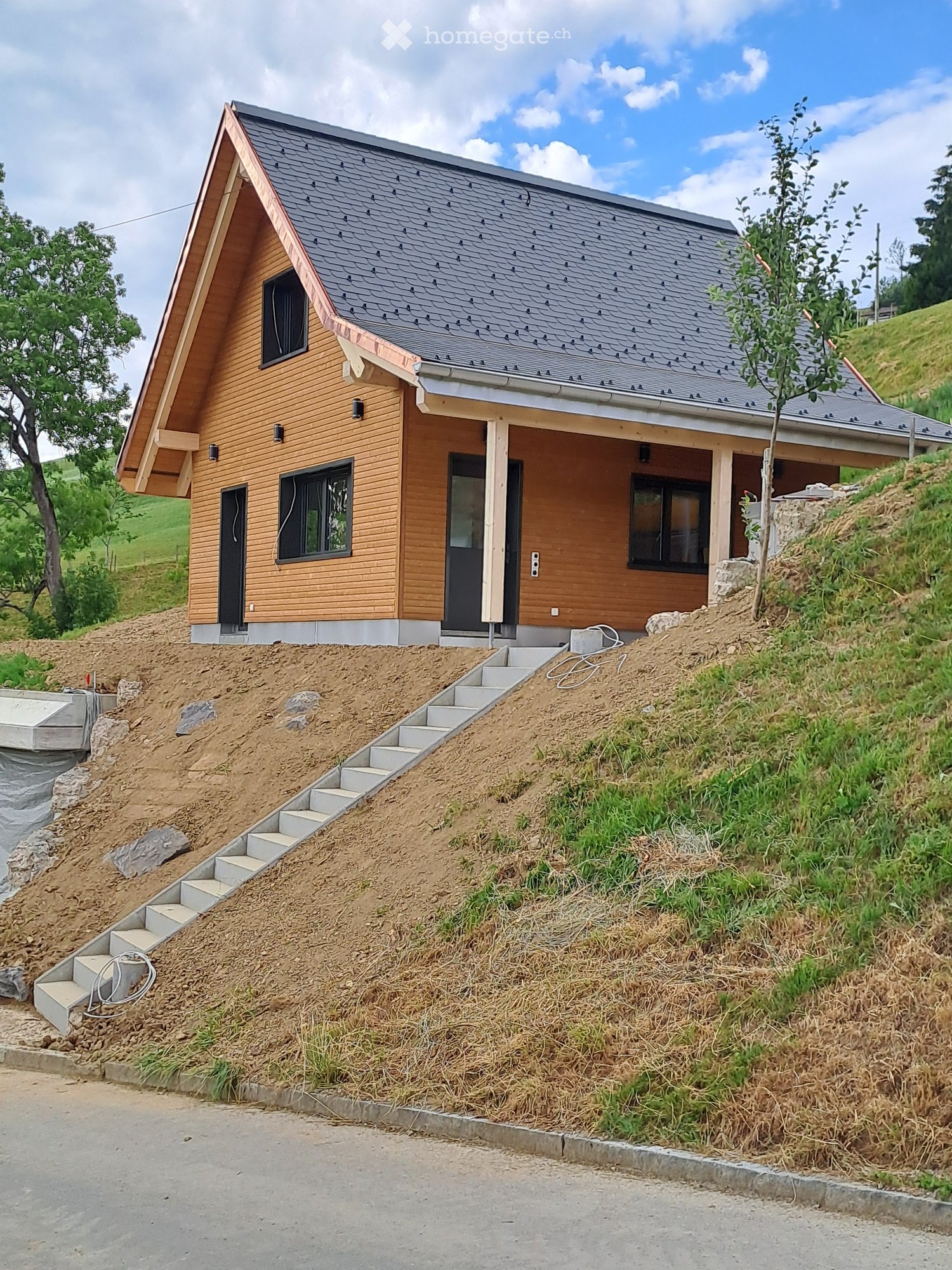 Modern two-story house with wooden siding, grey roof, and raised ground floor accessed by concrete steps.