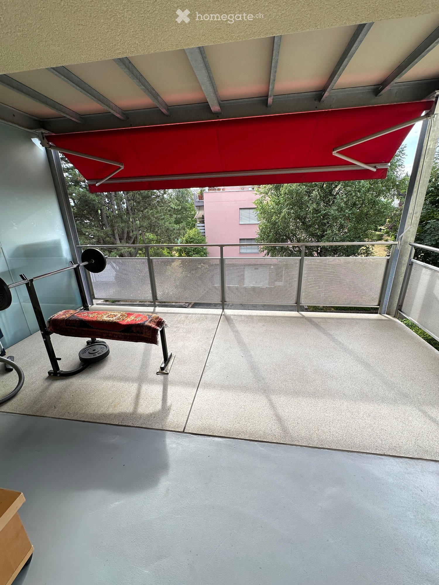Balcony with a red awning, metal railing, and a small bench with a patterned cushion.